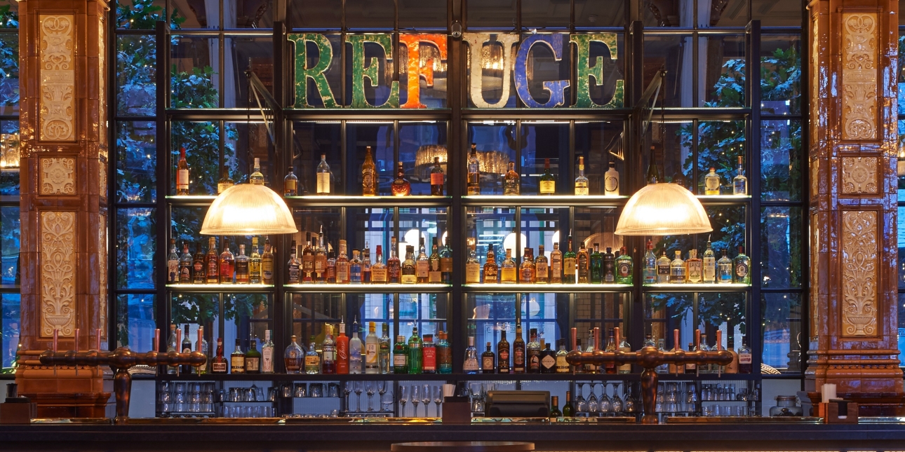 A bar with two art deco lamps hanging from the ceiling above with lots of bottles on the shelves with coloured letters spelling out REFUGE at the top